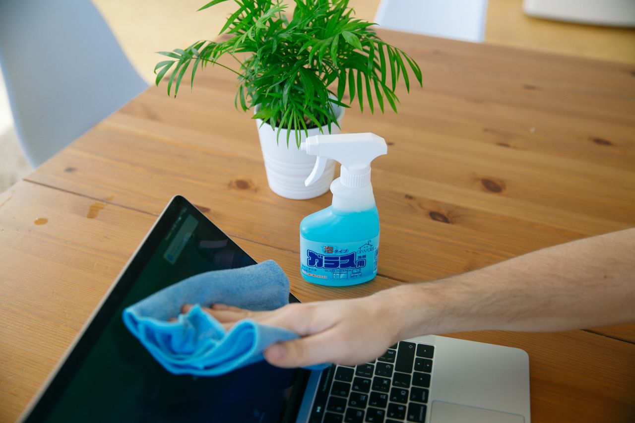 Gros plan d’une main nettoyant l’écran d’un ordinateur portable avec un chiffon bleu et un vaporisateur sur un bureau en bois.